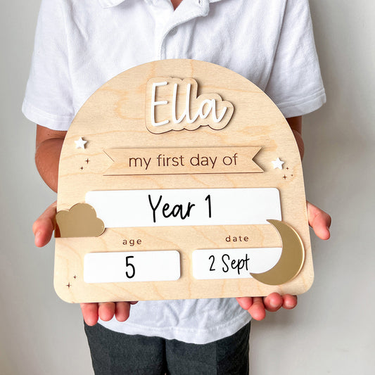 Wooden plaque with 'Ella' and details of a child's first day of school held by a person.