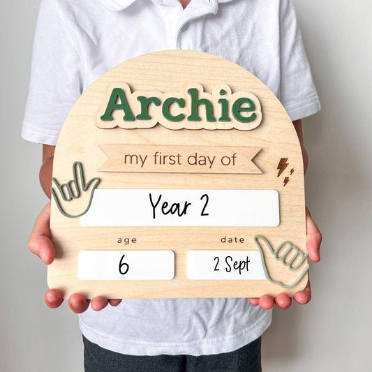 Wooden sign with 'Archie' and 'my first day of Year 2' text held by a person.