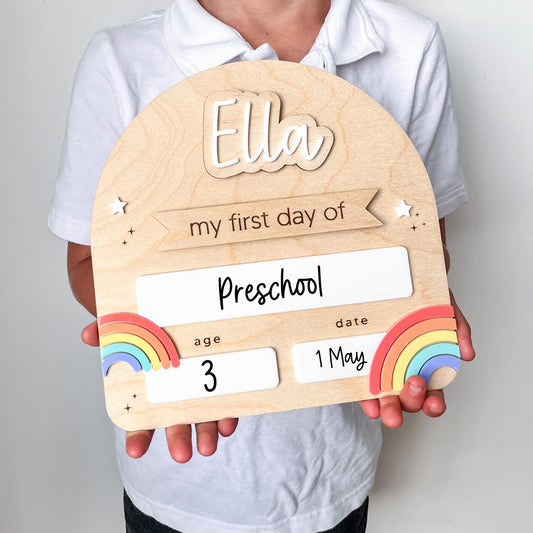 Wooden plaque with 'Ella' and 'my first day of preschool' text, held by a person.