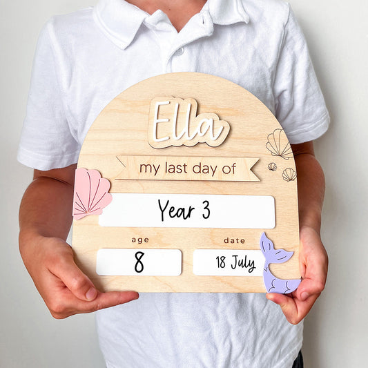 Person holding a wooden plaque with 'Ella' and 'my last day of Year 3' written on it.