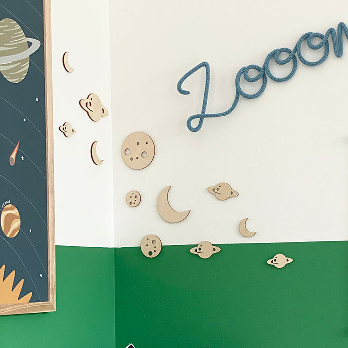 A styled children's bedroom corner with a green and white painted wall, wooden moon and planet wall decals, a blue 'Loom' wall sign, a framed 'To The Moon!' poster, and plush pillows shaped like a rocket ship and a space capsule on a bed.