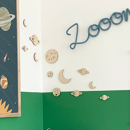 A styled children's bedroom corner with a green and white painted wall, wooden moon and planet wall decals, a blue 'Loom' wall sign, a framed 'To The Moon!' poster, and plush pillows shaped like a rocket ship and a space capsule on a bed.