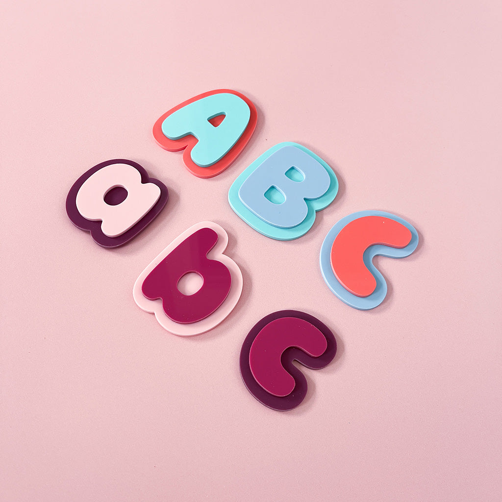 Six colorful alphabet letters arranged on a soft pink background, including A, B, C, a, b, and c in pastel and bright shades of blue, pink, purple, and coral.
