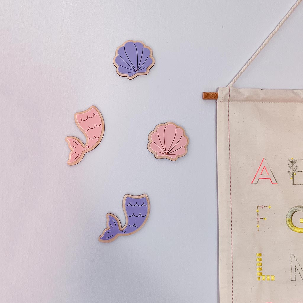 Four mermaid-themed wooden decor, including two purple mermaid tails and two pink seashells, arranged on a white wall next to a canvas banner.