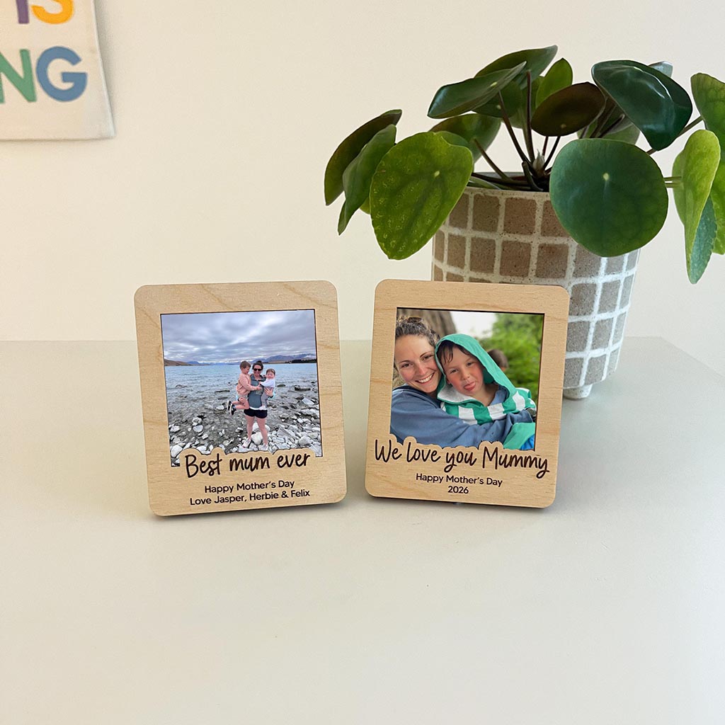 Two wooden photo frames with engraved text and family photos on a light surface with a plant in the background.
