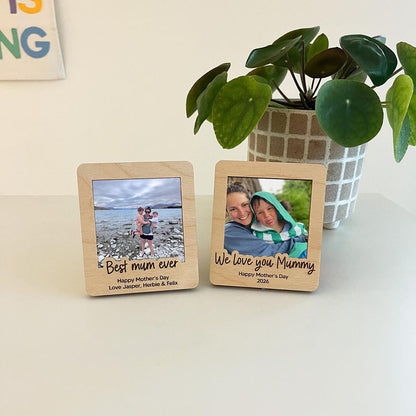 Two wooden photo frames with engraved text and family photos on a light surface with a plant in the background.