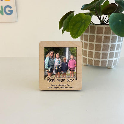 Wooden photo frame with a family photo and 'Best mum ever' text, placed on a surface with a plant in the background.