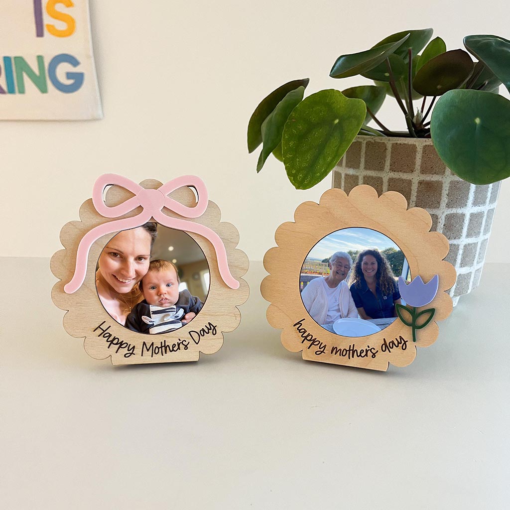 Two wooden photo frames with 'Happy Mother's Day' text, one with a pink bow and the other with a purple flower, on a light surface with a plant in the background.