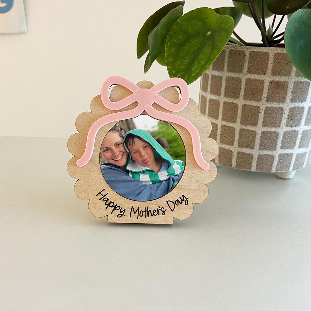 Wooden photo frame with pink bow and 'Happy Mother's Day' text, featuring a photo of a woman and child, on a light surface with a plant in the background.