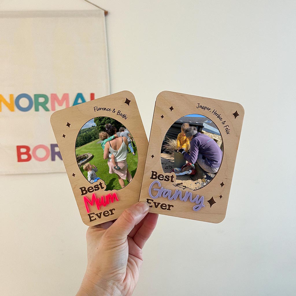 Two wooden frames with photos and text held by a hand, with a colourful 'NORMAL' sign in the background.