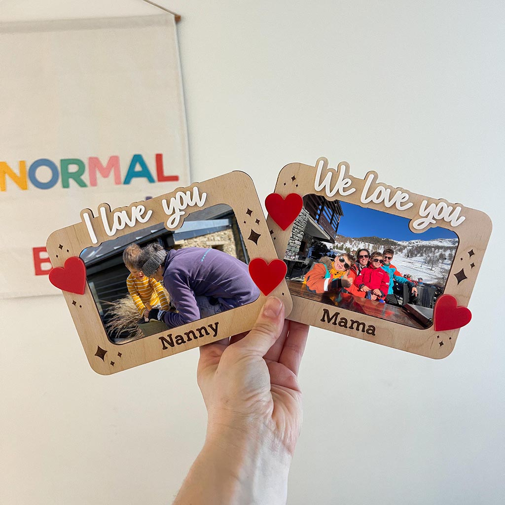 Two wooden photo frames with hearts and text, held by a hand against a light background.