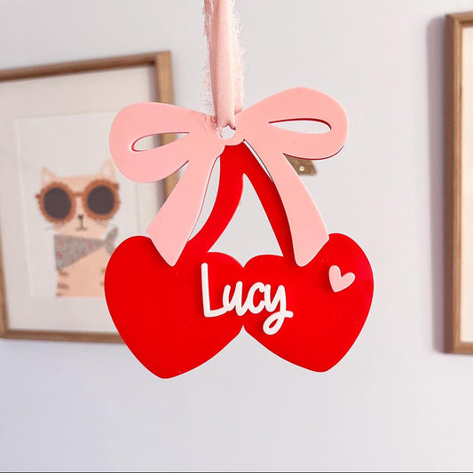 Red heart-shaped ornament with a pink bow and 'Lucy' written on it, held by a hand against a light background.
