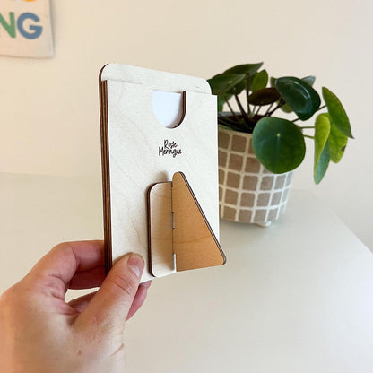 Hand holding a wooden phone stand with 'Rose Manque' branding, plant in the background