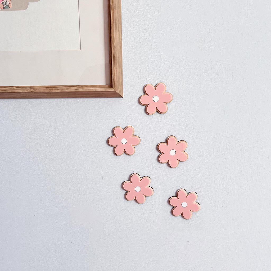Five pink flower-shaped wall hooks with white centers, arranged on a light gray wall next to a wooden picture frame.