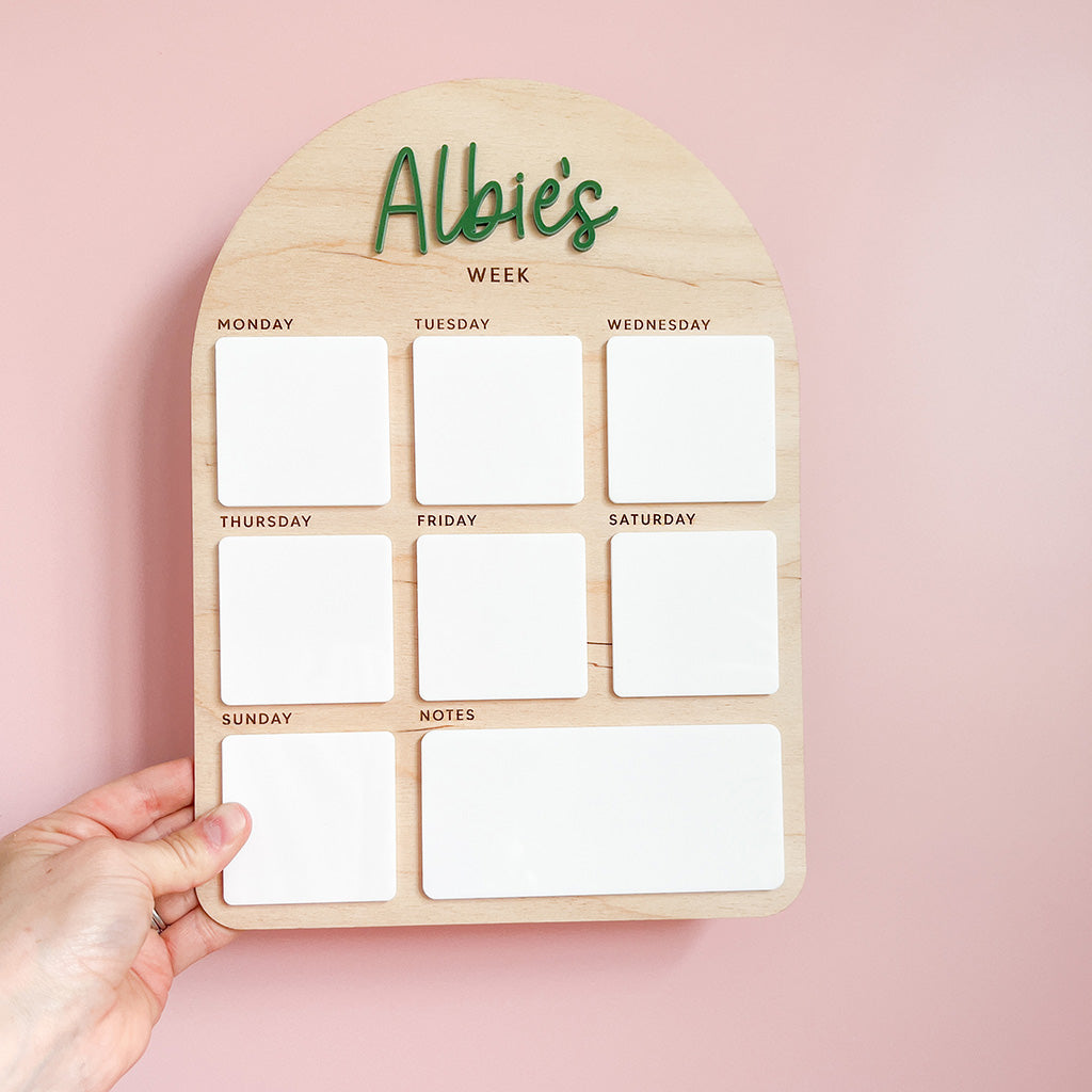 A wooden weekly planner board with the text 'Albie's WEEK' at the top, featuring seven white magnetic slots for Monday through Sunday and a larger white notes section. A hand is holding the board against a soft pink background.