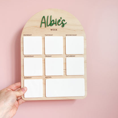 A wooden weekly planner board with the text 'Albie's WEEK' at the top, featuring seven white magnetic slots for Monday through Sunday and a larger white notes section. A hand is holding the board against a soft pink background.